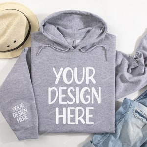 Pode incluir: Uma camisa de moletom cinza com capuz com o texto "YOUR DESIGN HERE" impresso em branco na frente. A camisa de moletom est&aacute; colocada sobre uma superf&iacute;cie branca com um chap&eacute;u de palha e uma cal&ccedil;a jeans azul.