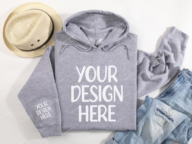 Pode incluir: Uma camisa de moletom cinza com capuz com o texto "YOUR DESIGN HERE" impresso em branco na frente. A camisa de moletom est&aacute; colocada sobre uma superf&iacute;cie branca com um chap&eacute;u de palha e uma cal&ccedil;a jeans azul.