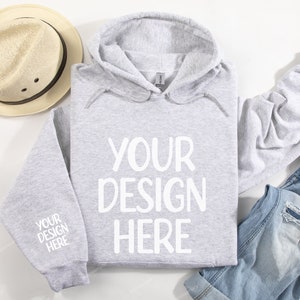 Pode incluir: Uma camisola de capuz cinzenta com o texto "YOUR DESIGN HERE" impresso a branco na frente. A camisola est&aacute; colocada sobre uma superf&iacute;cie branca com um chap&eacute;u de palha e cal&ccedil;as de ganga azuis.
