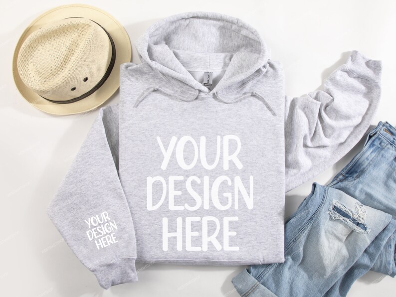 Pode incluir: Uma camisola de capuz cinzenta com o texto "YOUR DESIGN HERE" impresso a branco na frente. A camisola est&aacute; colocada sobre uma superf&iacute;cie branca com um chap&eacute;u de palha e cal&ccedil;as de ganga azuis.