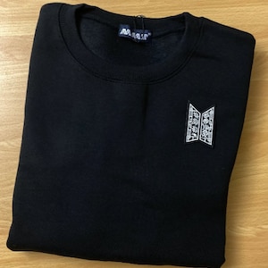 Puede incluir: Sudadera negra con un logotipo bordado en blanco que presenta las letras "BTS" y texto en coreano. El logotipo está en el lado izquierdo del pecho.
