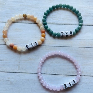 May include: Three beaded bracelets with white number tiles. The first bracelet is made of light brown and white beads and has the number "511" on it. The second bracelet is made of green beads and has the number "325" on it. The third bracelet is made of pink beads and has the number "211" on it.