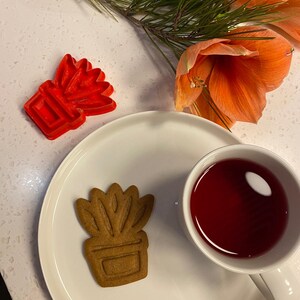 Peut inclure: Un biscuit en pain d'épices en forme de plante en pot est posé sur une assiette blanche, à côté d'une tasse de liquide rouge. Un emporte-pièce rouge en forme de plante et des fleurs orange sont également dans l'image.
