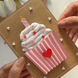 May include: A handmade birthday card with a cupcake design. The cupcake has white frosting with red and pink heart decorations, a pink paper wrapper, and a candle. The card is brown with the word "Birthday" and "DIY CUPCAKE CARD" written on it.
