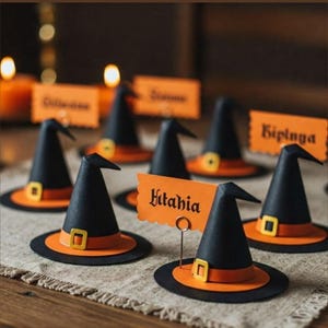 Puede incluir: Una colección de miniaturas de sombreros de bruja para tarjetas de mesa. Cada sombrero es negro con un borde naranja y una hebilla dorada. Tarjetas de nombre naranjas se insertan en los sombreros. La palabra "Kitahia" es visible en una tarjeta.
