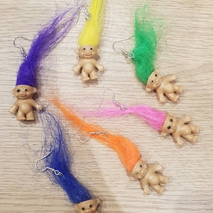 May include: Six pairs of troll doll earrings with colourful hair. Each earring features a small troll doll with a different coloured hairpiece, including purple, yellow, green, blue, pink, and orange.