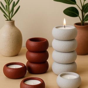 May include: A collection of decorative candles in various sizes and colors. The candles are in a textured, round shape, with a stack of four gray candles and several individual red-brown candles. A lit candle is in the center of the gray stack. Plants in vases are in the background.