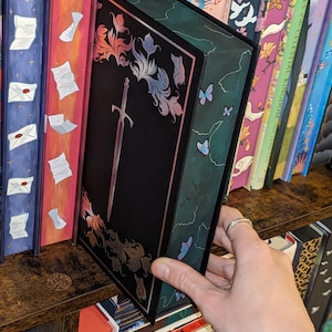 Puede incluir: Un libro negro con un dise&ntilde;o de espada plateada y detalles florales. El libro est&aacute; en una estanter&iacute;a de madera con otros libros. El lomo del libro tiene un fondo verde con mariposas azules. El libro es parte de una colecci&oacute;n.