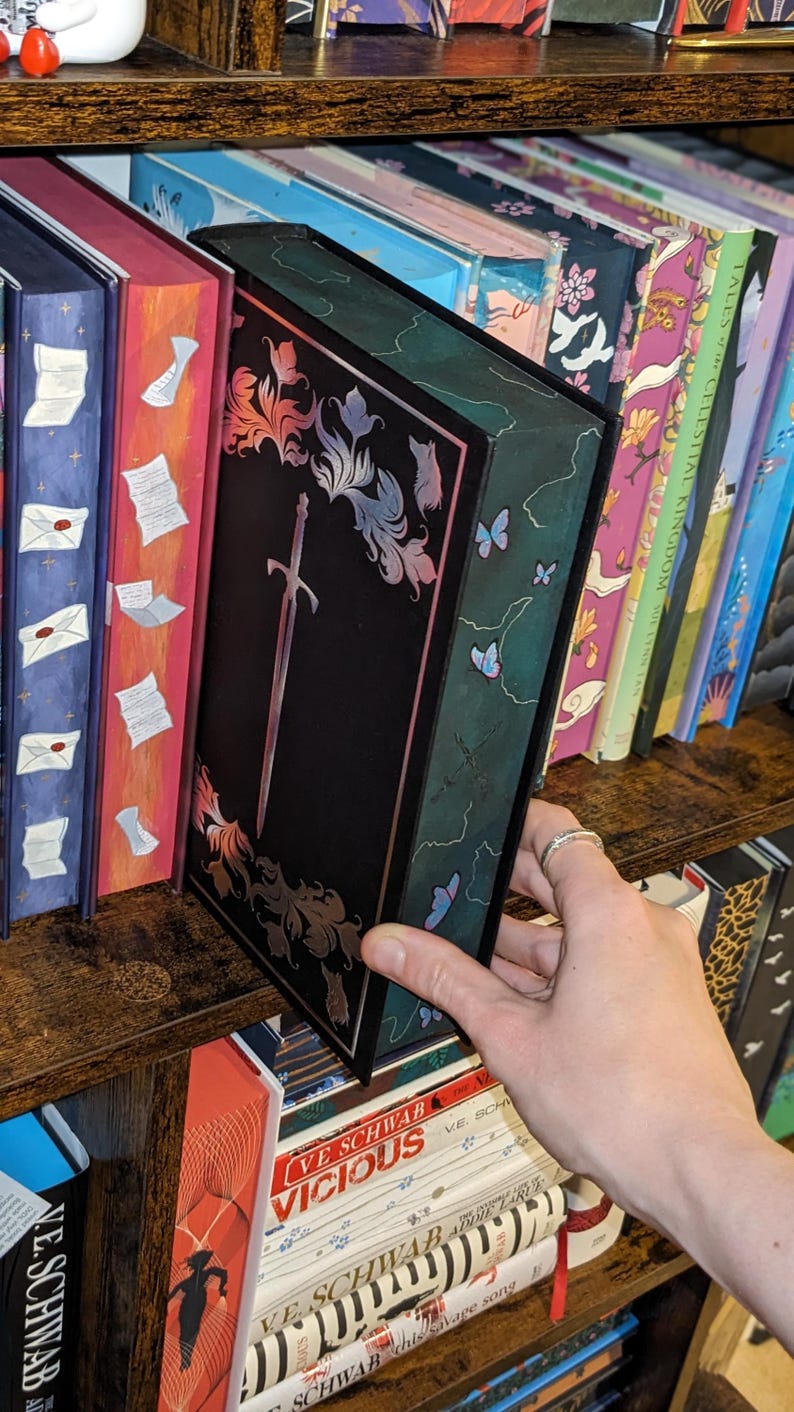 Puede incluir: Un libro negro con un dise&ntilde;o de espada plateada y detalles florales. El libro est&aacute; en una estanter&iacute;a de madera con otros libros. El lomo del libro tiene un fondo verde con mariposas azules. El libro es parte de una colecci&oacute;n.
