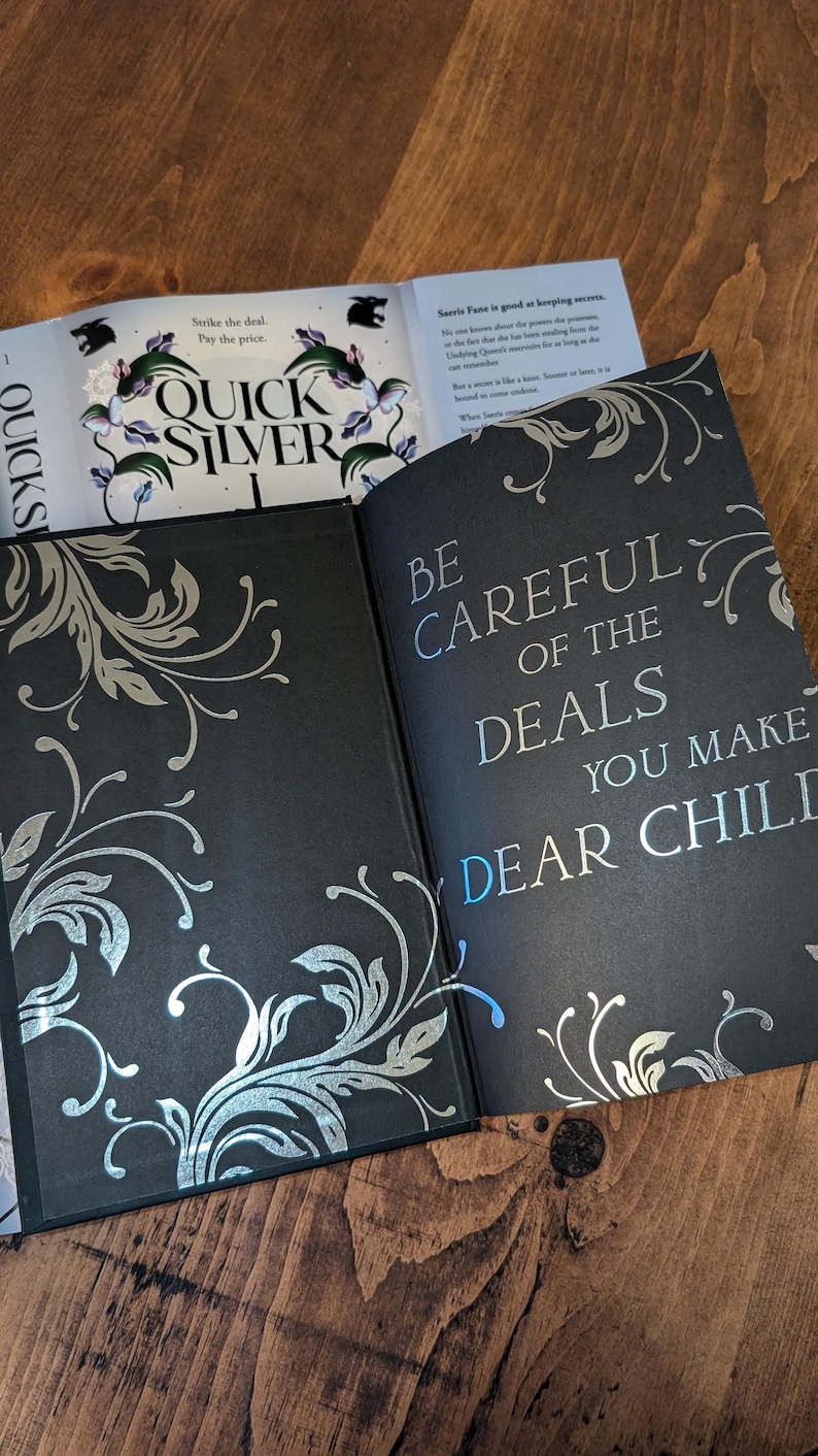 Puede incluir: Libro abierto con p&aacute;ginas negras y dise&ntilde;os florales plateados. El texto "QUICK SILVER" es visible en una p&aacute;gina, y "BE CAREFUL OF THE DEALS YOU MAKE DEAR CHILD" en otra. El libro est&aacute; sobre una superficie de madera.