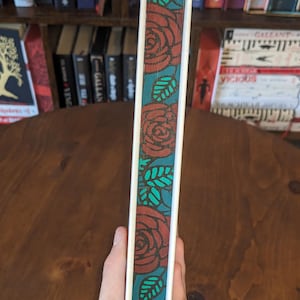 Puede incluir: Un diario blanco con un dise&ntilde;o floral rojo y turquesa en el lomo. El diario est&aacute; sostenido en una mano con una pulsera de plata.