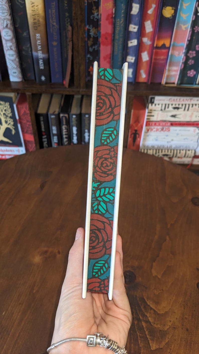 Puede incluir: Un diario blanco con un dise&ntilde;o floral rojo y turquesa en el lomo. El diario est&aacute; sostenido en una mano con una pulsera de plata.