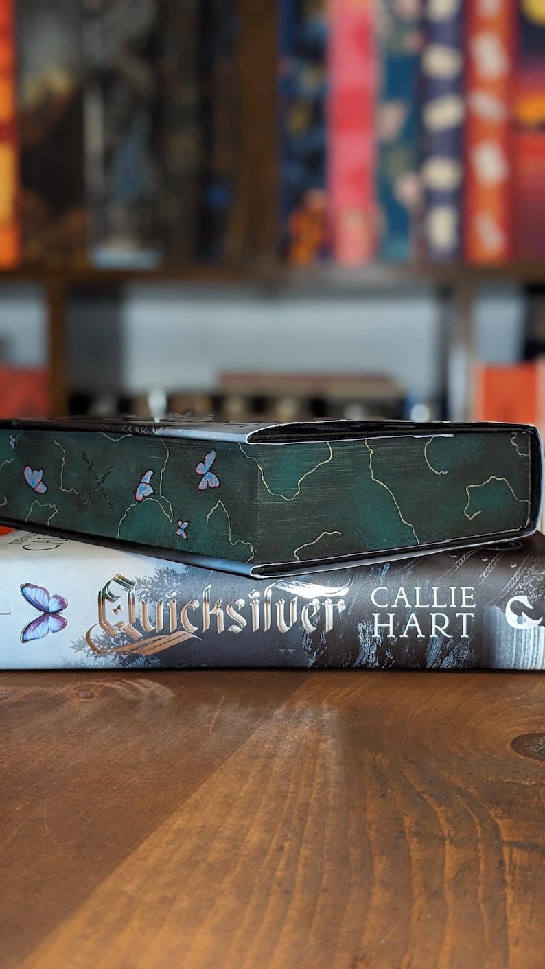 Puede incluir: Una pila de libros. El libro superior tiene una cubierta verde oscuro con ilustraciones de mariposas. El libro de abajo se titula "Quicksilver" de Callie Hart. Los libros est&aacute;n sobre una superficie de madera, con un fondo borroso de otros libros.