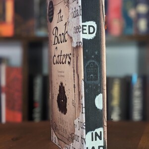 May include: A book titled "The Book Eaters" with a brown and white cover. The book is on a wooden shelf with other books behind it. The spine of the book has the words "TH IN AP ED" on it.
