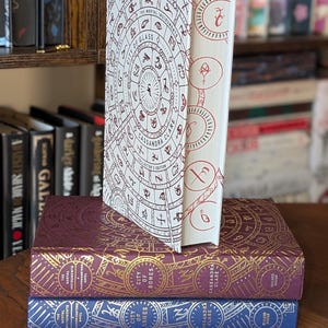 Könnte beinhalten: Ein Stapel von drei gebundenen Büchern mit aufwendigen Designs. Das oberste Buch ist weiß mit einem roten und schwarzen Kreisdesign und dem Text "The Mortal Instruments City of Glass Cassandra Clare Collector's Edition". Das mittlere Buch ist burgunderrot mit goldenen Akzenten und dem Text "The Mortal Instruments City of Bones Cassandra Clare". Das unterste Buch ist blau mit goldenen Akzenten und dem Text "The Mortal Instruments City of Ashes Cassandra Clare".