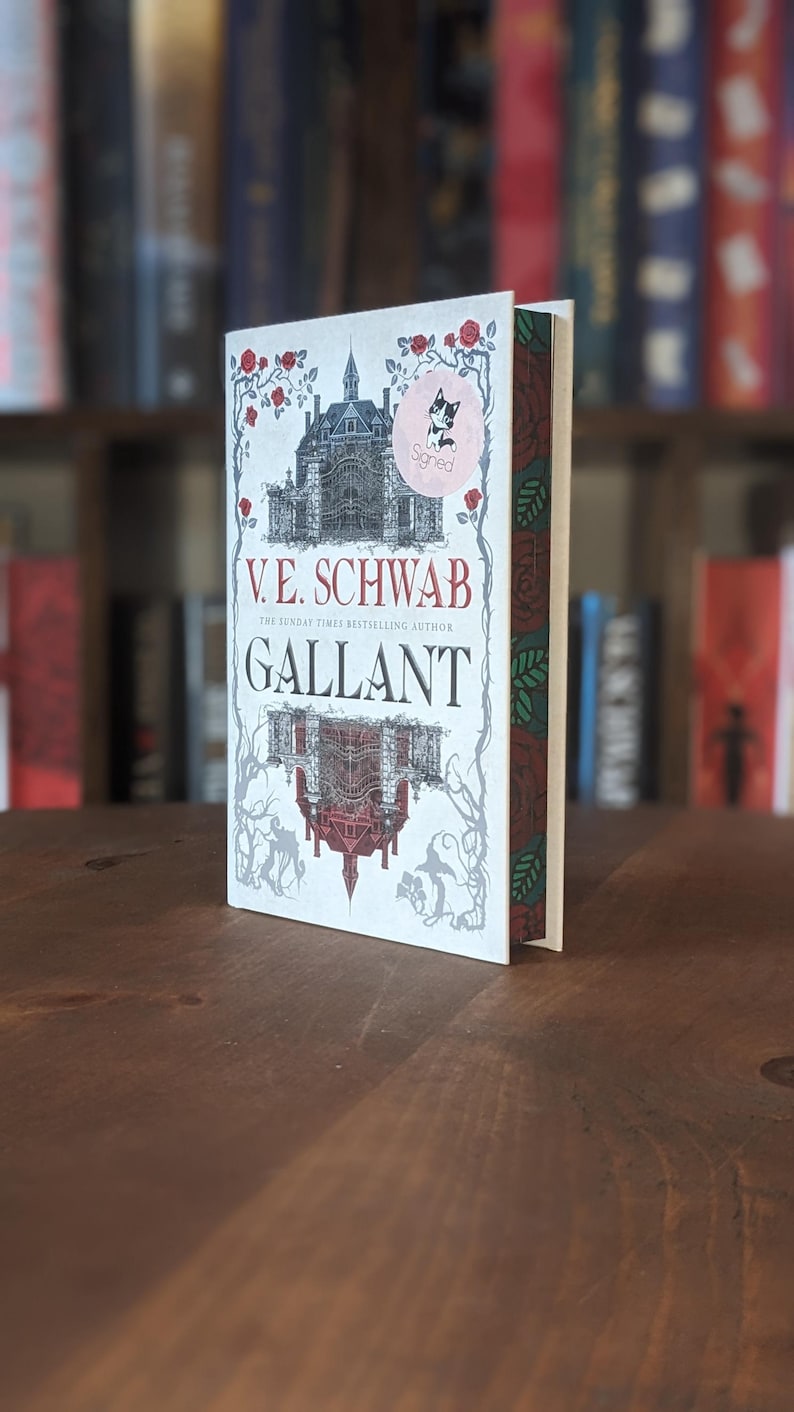 Puede incluir: Un libro de tapa dura blanco con una ilustraci&oacute;n en blanco y negro de una casa en la portada. El t&iacute;tulo del libro es "Gallant" de V.E. Schwab, la autora m&aacute;s vendida del Sunday Times. El libro tiene un dise&ntilde;o floral rojo y verde en el lomo.