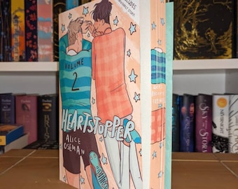 Könnte beinhalten: Band 2 des Graphic Novels Heartstopper von Alice Oseman. Das Cover zeigt zwei junge Männer, einer trägt ein blaues und weißes Rugby-Trikot und der andere ein rosa und weißes Hemd. Der Hintergrund ist hellblau mit weißen Sternen.