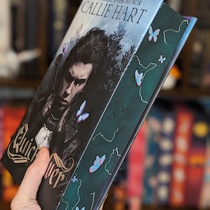 Puede incluir: Un libro titulado "Quick Silver" de Callie Hart. La portada presenta una ilustraci&oacute;n oscura de una persona con una espada, rodeada de mariposas. El lomo es verde oscuro con detalles de mariposas y el texto "Quick Silver".