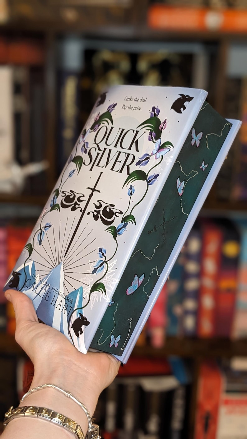 Puede incluir: Un libro de tapa dura titulado "QUICKSILVER" de Callie Hart. La portada es azul claro con un dise&ntilde;o de espada, detalles florales y el texto "Strike the deal. Pay the price." El lomo del libro es verde oscuro con ilustraciones de mariposas.