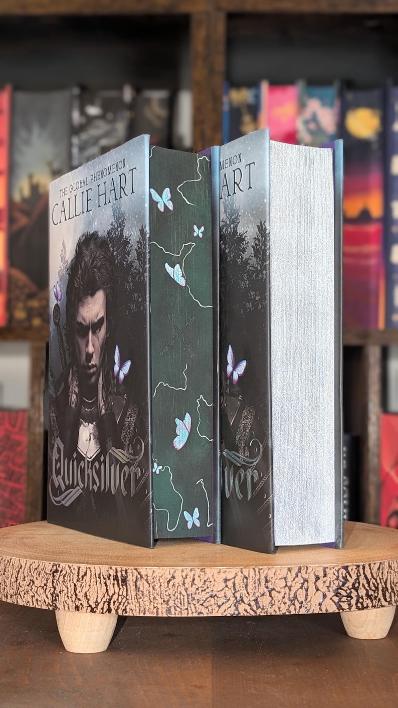 Puede incluir: Tres libros de tapa dura con el t&iacute;tulo "Quicksilver" de Callie Hart. Las portadas de los libros presentan una combinaci&oacute;n de colores oscuros con ilustraciones de una persona, mariposas y texto. Los libros se exhiben en un soporte de madera.