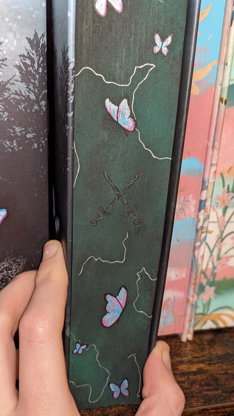 Puede incluir: Lomo de libro verde oscuro con un dise&ntilde;o de mariposas rosas y azules, con contornos blancos. Tambi&eacute;n se representan dos espadas cruzadas. El lomo est&aacute; sostenido por una mano, con otros libros visibles en el fondo.