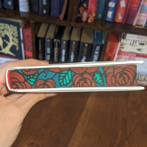 Puede incluir: Un libro blanco con un dise&ntilde;o floral rojo y turquesa en el lomo. El libro est&aacute; siendo sostenido por una mano.
