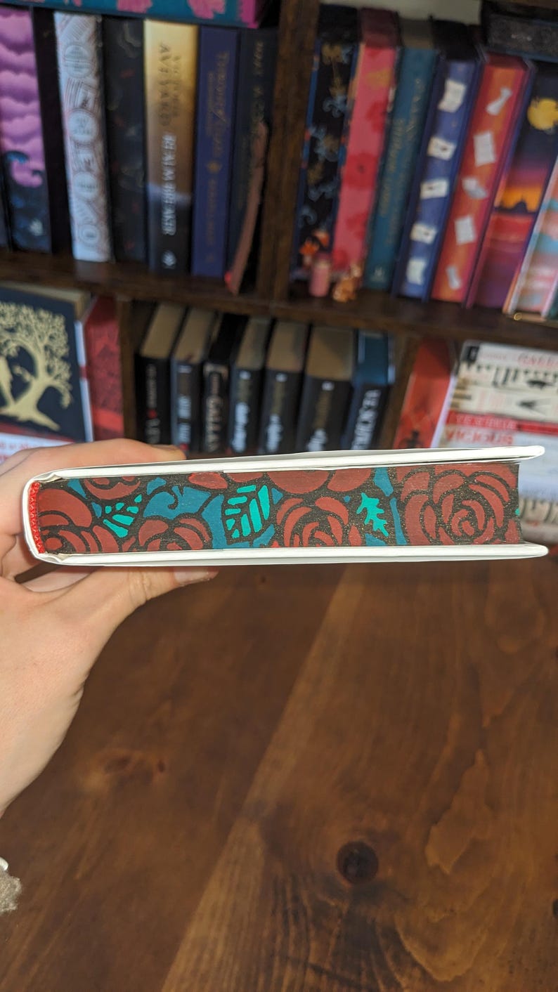 Puede incluir: Un libro blanco con un dise&ntilde;o floral rojo y turquesa en el lomo. El libro est&aacute; siendo sostenido por una mano.