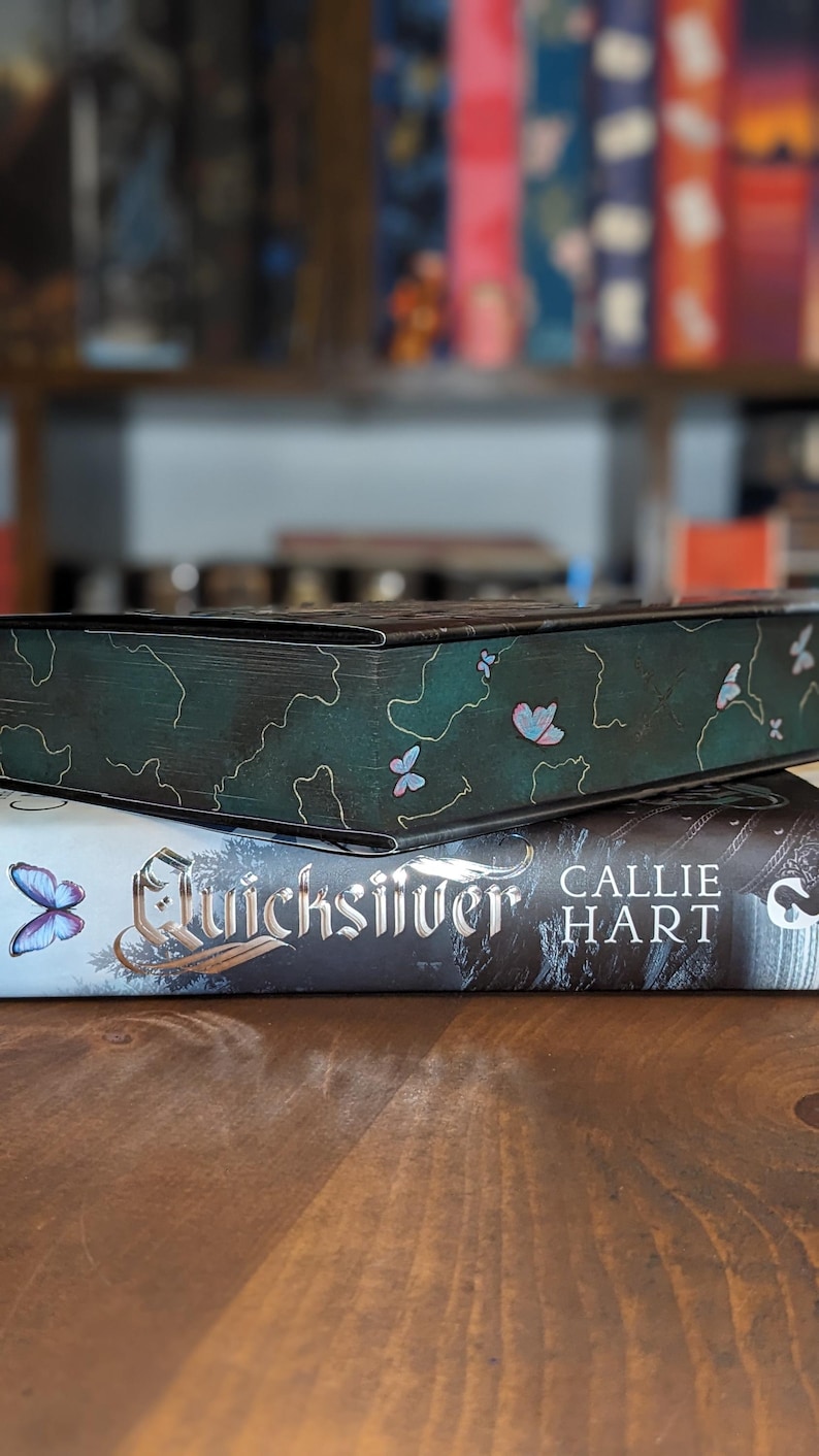 Puede incluir: Dos libros apilados sobre una superficie de madera. El libro superior es de color verde oscuro con un dise&ntilde;o de mariposa. El libro inferior se titula "Quicksilver" de Callie Hart. Libros frente a una estanter&iacute;a borrosa.