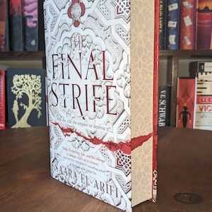 Puede incluir: Un libro blanco con un diseño rojo y dorado en la portada. El título "The Final Strife" está en letras rojas. El libro se titula "The Final Strife" y el subtítulo es "Will she rule the empire or destroy it?" El autor es Saara El-Arifi.
