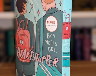 Könnte beinhalten: Ein Cover eines Graphic Novels in den Farben Blau, Grün und Pink. Das Cover zeigt zwei Jungen, die nebeneinander gehen, einer mit einem roten Rucksack und der andere mit einer blauen Tasche. Der Titel "Heartstopper" ist in großen, pinkfarbenen Buchstaben geschrieben. Der Text "Boy Meets Boy" ist in kleineren Buchstaben unter dem Titel geschrieben. Der Text "Alice Oseman" ist in kleineren Buchstaben unten auf dem Cover geschrieben. Der Text "Netflix Coming to Netflix" ist in einem Kreis auf dem Cover geschrieben.