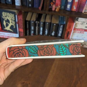 Puede incluir: Primer plano del lomo de un libro con un dise&ntilde;o floral rojo, blanco y turquesa. El dise&ntilde;o presenta rosas y hojas.