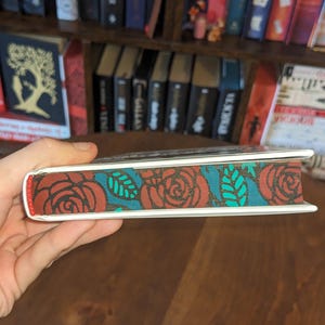 Puede incluir: Primer plano del lomo de un libro con un dise&ntilde;o floral rojo, blanco y turquesa. El dise&ntilde;o presenta rosas y hojas.