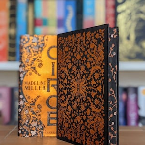 Könnte beinhalten: Zwei gebundene Bücher mit goldenen und schwarzen floralen Mustern auf den Umschlägen. Das Buch links trägt den Titel "Circe" und der Autor "Madeline Miller" ist auf dem Umschlag gedruckt. Das Buch rechts hat ein komplexeres Blumenmuster.