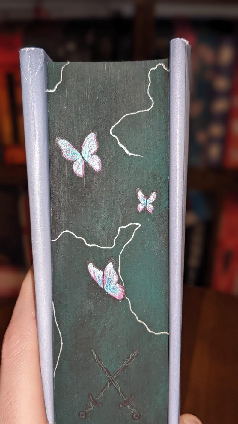 Puede incluir: Lomo de libro con fondo verde oscuro, con tres mariposas de alas rosas y azules y arte lineal blanco. Dos espadas cruzadas en la parte inferior. Los bordes son de color gris claro.