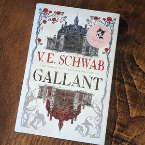 Puede incluir: Una cubierta de libro blanca con una ilustraci&oacute;n en blanco y negro de una casa con una torre de reloj. El t&iacute;tulo "V.E. SCHWAB" y "GALLANT" est&aacute;n impresos en negro y gris. El libro est&aacute; firmado con una pegatina que dice "Firmado" y tiene una ilustraci&oacute;n en blanco y negro de un gato.