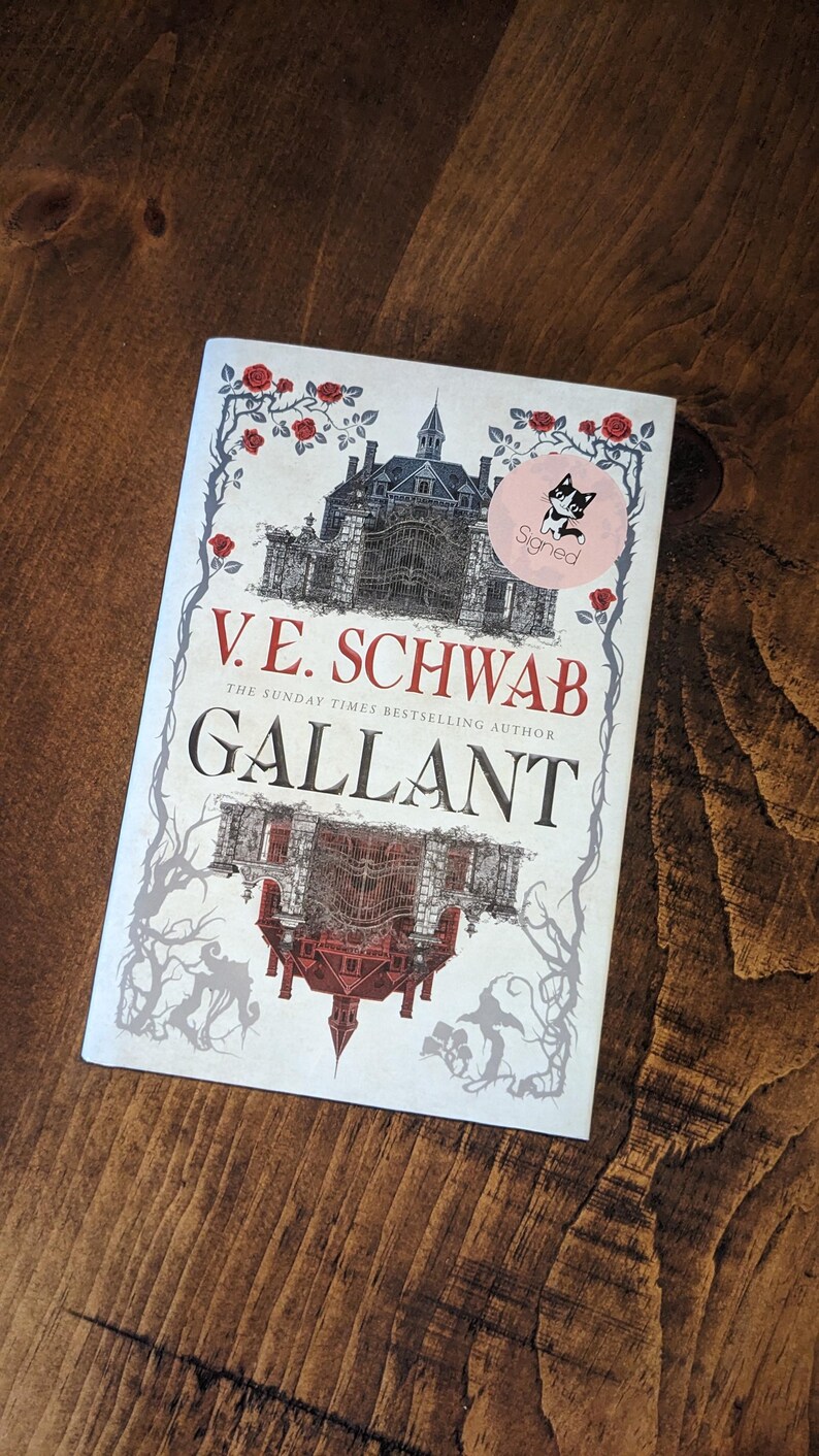 Puede incluir: Una cubierta de libro blanca con una ilustraci&oacute;n en blanco y negro de una casa con una torre de reloj. El t&iacute;tulo "V.E. SCHWAB" y "GALLANT" est&aacute;n impresos en negro y gris. El libro est&aacute; firmado con una pegatina que dice "Firmado" y tiene una ilustraci&oacute;n en blanco y negro de un gato.