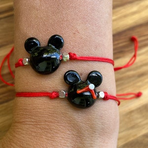 K&ouml;nnte beinhalten: Zwei rote Armb&auml;nder mit schwarzen Glas-Mickey-Mouse-Anh&auml;ngern. Die Anh&auml;nger haben silberne Akzente.