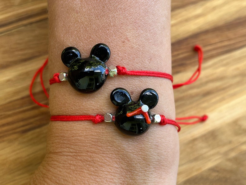 K&ouml;nnte beinhalten: Zwei rote Armb&auml;nder mit schwarzen Glas-Mickey-Mouse-Anh&auml;ngern. Die Anh&auml;nger haben silberne Akzente.