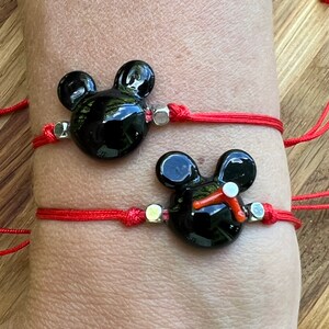 K&ouml;nnte beinhalten: Zwei rote Armb&auml;nder mit schwarzen Glas-Mickey-Mouse-Anh&auml;ngern. Die Anh&auml;nger haben silberne Akzente.