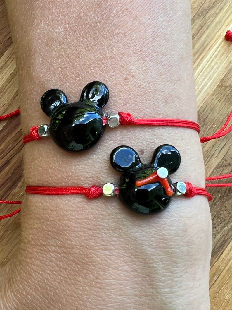 K&ouml;nnte beinhalten: Zwei rote Armb&auml;nder mit schwarzen Glas-Mickey-Mouse-Anh&auml;ngern. Die Anh&auml;nger haben silberne Akzente.