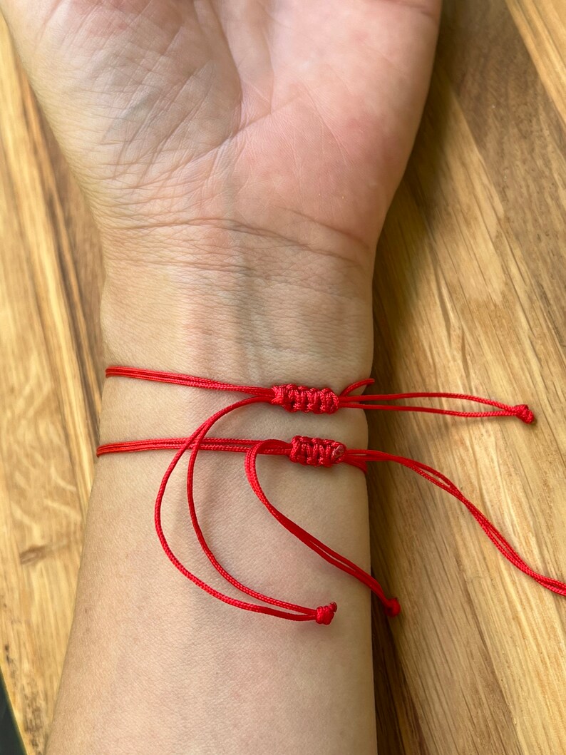 K&ouml;nnte beinhalten: Drei rote Armb&auml;nder aus Schnur, die um ein Handgelenk gebunden sind. Die Armb&auml;nder bestehen aus d&uuml;nner roter Schnur und haben Knoten an den Enden.