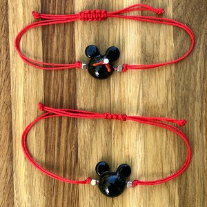 K&ouml;nnte beinhalten: Zwei rote Armb&auml;nder aus Schnur mit schwarzen Mickey-Maus-Anh&auml;ngern. Die Anh&auml;nger haben wei&szlig;e Akzente und sind mit silbernen Perlen an den Armb&auml;ndern befestigt.