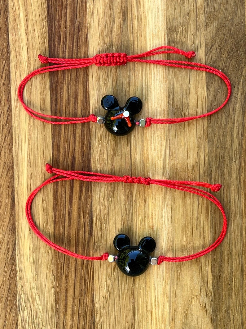 K&ouml;nnte beinhalten: Zwei rote Armb&auml;nder aus Schnur mit schwarzen Mickey-Maus-Anh&auml;ngern. Die Anh&auml;nger haben wei&szlig;e Akzente und sind mit silbernen Perlen an den Armb&auml;ndern befestigt.