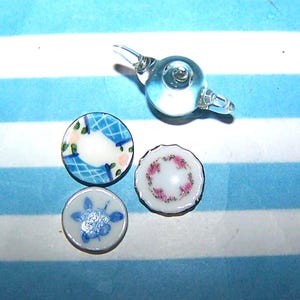 May include: Miniature glass teapot and three small decorative plates. The plates feature floral and geometric patterns in blue, pink, and white. The teapot is clear glass.
