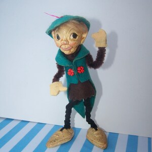 May include: A vintage elf doll with a green felt hat, a green felt jacket with red flower accents, brown felt pants, and black pipe cleaner arms and legs. The doll is wearing brown felt shoes.