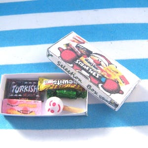Può includere: Una scatola rettangolare in miniatura di cartone bianco con l'etichetta Selection Box e una grafica colorata di un personaggio di Babbo Natale. All'interno della scatola aperta ci sono piccole repliche giocattolo di dolciumi e un lecca-lecca.