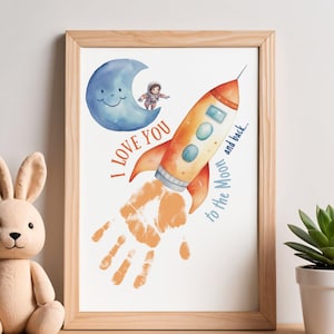 May include: A framed print featuring a watercolor illustration of a rocket ship blasting off, with a handprint trail. The print includes a blue crescent moon with a smiling face and an astronaut. Text reads "I love you to the moon and back..."