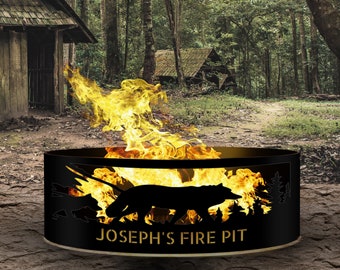 Op de afbeelding: Een zwarte metalen vuurplaats met een wolf-silhouetontwerp en de tekst "JOSEPH'S FIRE PIT". De vuurplaats is omringd door vlammen en staat in een bosrijke omgeving.