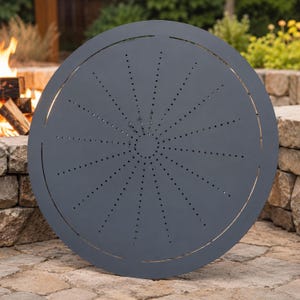 May include: A circular dark gray metal fire pit cover featuring a central solid disc surrounded by radiating lines of small, evenly spaced perforations. The outer edge of the disc has a thin, circular cut-out detail along the perimeter.
