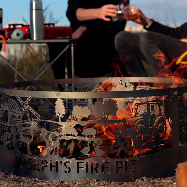 K&ouml;nnte beinhalten: Ein schwarzer Metall-Feuerplatz mit einem ausgeschnittenen Design eines Traktors, B&auml;umen und den Worten "Joseph's Fire Pit". Der Feuerplatz ist von Flammen umgeben.